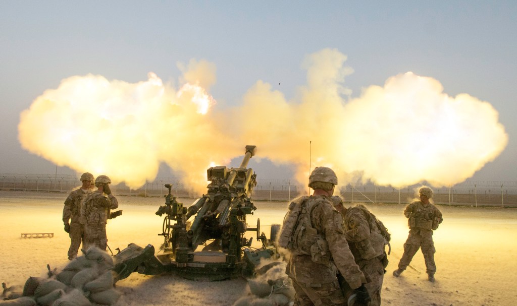 A towed artillery canon fires with large plumes of smoke and flame in a dusty environment.